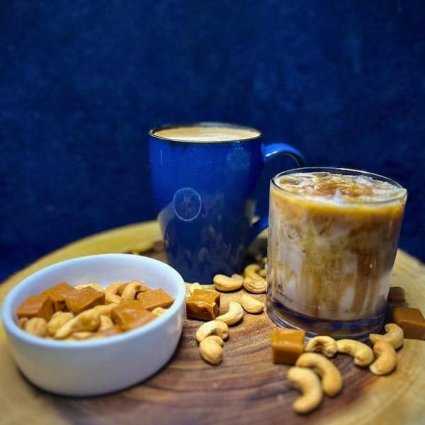 A blue mug of coffee, an iced coffee, cashews, and caramels on a wooden board.