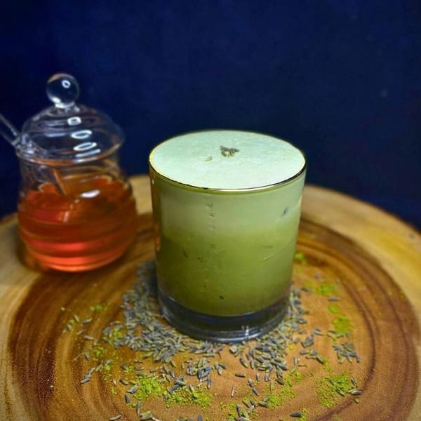 A green matcha latte with foam and lavender on a wooden board next to a honey jar.