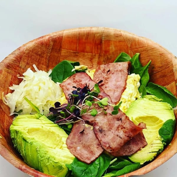 A breakfast bowl with spinach, avocado, bacon, sauerkraut, and microgreens in a wooden bowl.