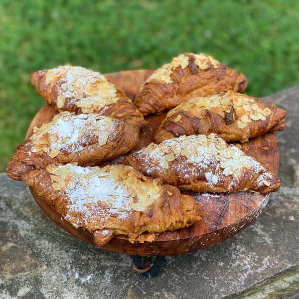 Six almond croissants with powdered sugar on a wooden platter, set against a green and stone background.