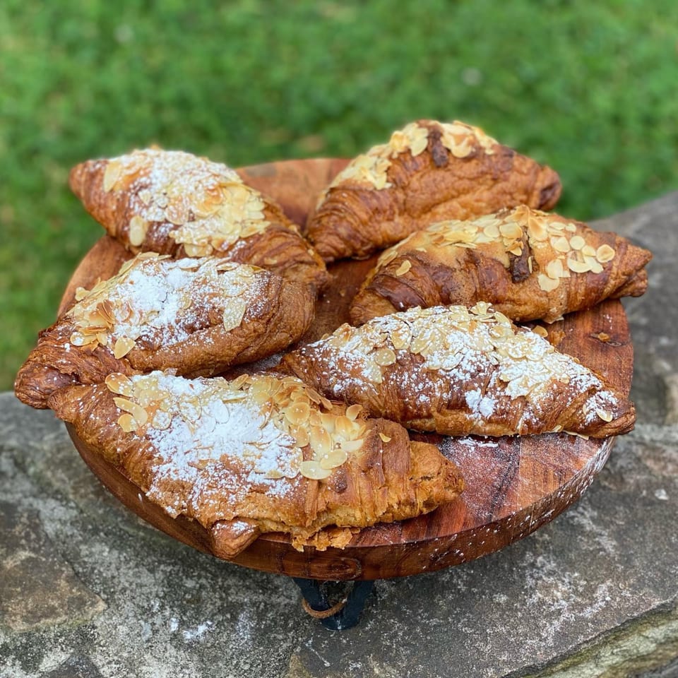 Six almond croissants with powdered sugar on a wooden platter, set against a green and stone background.